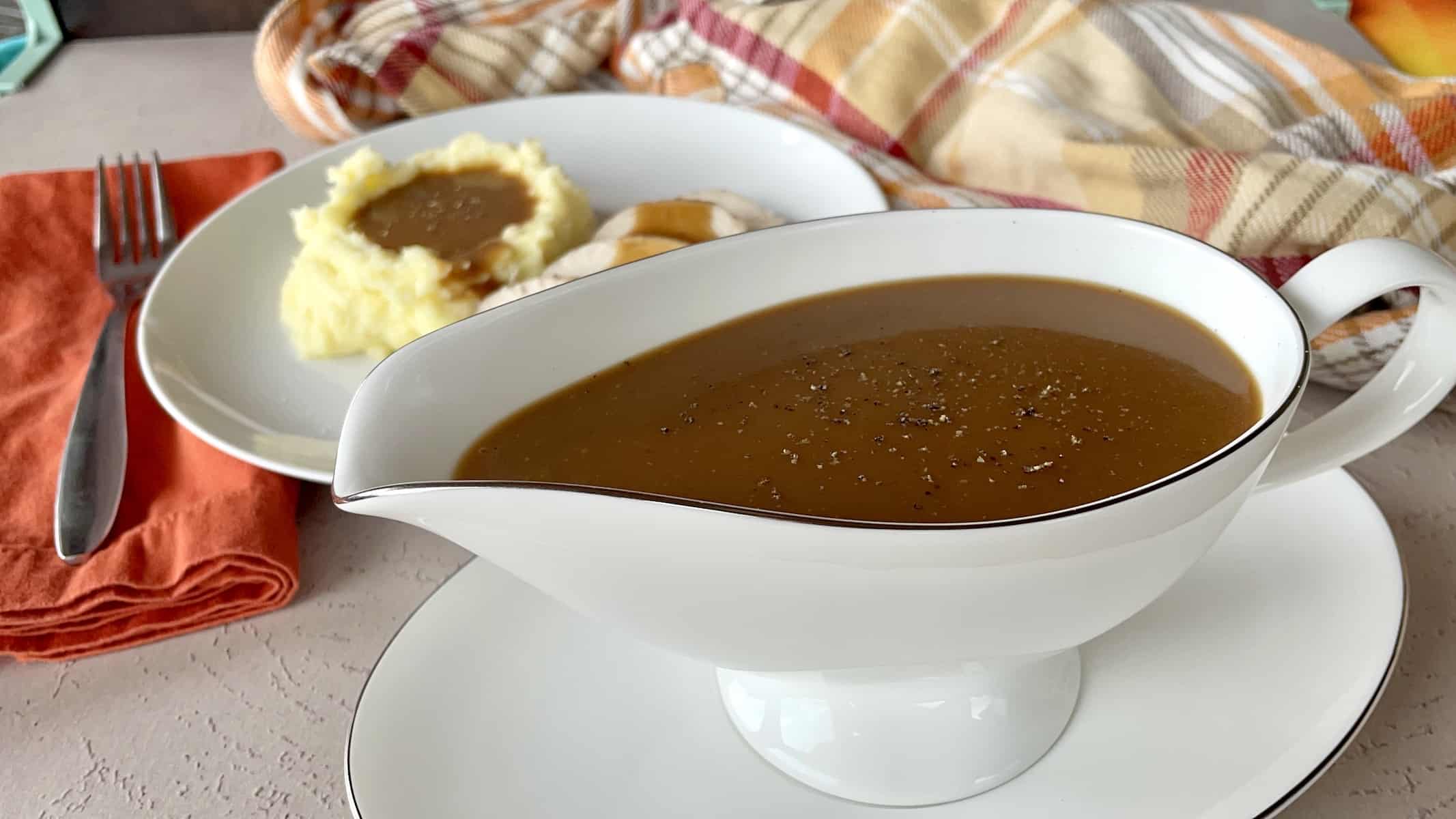 white gravy boat with brown gravy and a plate with served mashed potatoes and gravy next to a orange plaid apron