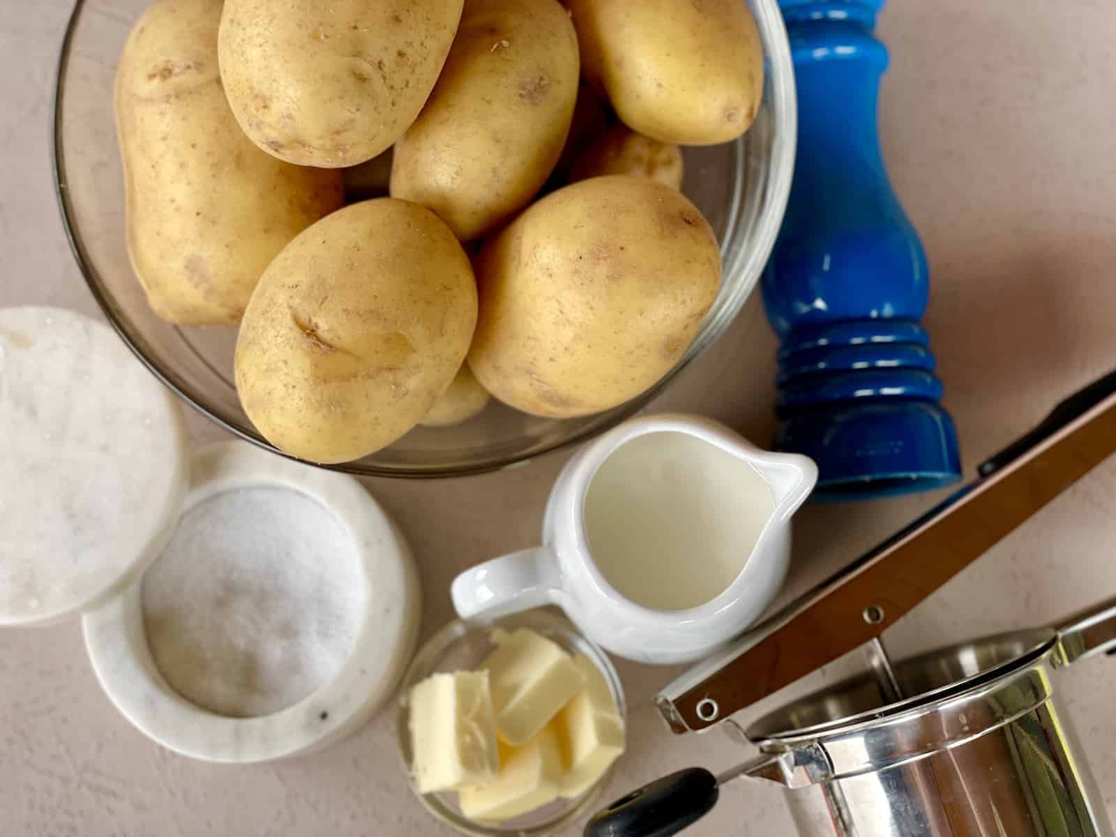 a bowl of yukon gold potatoes next to a salt jar cream in a white pitcher small bowl of butter a blue pepper grinder and ricer