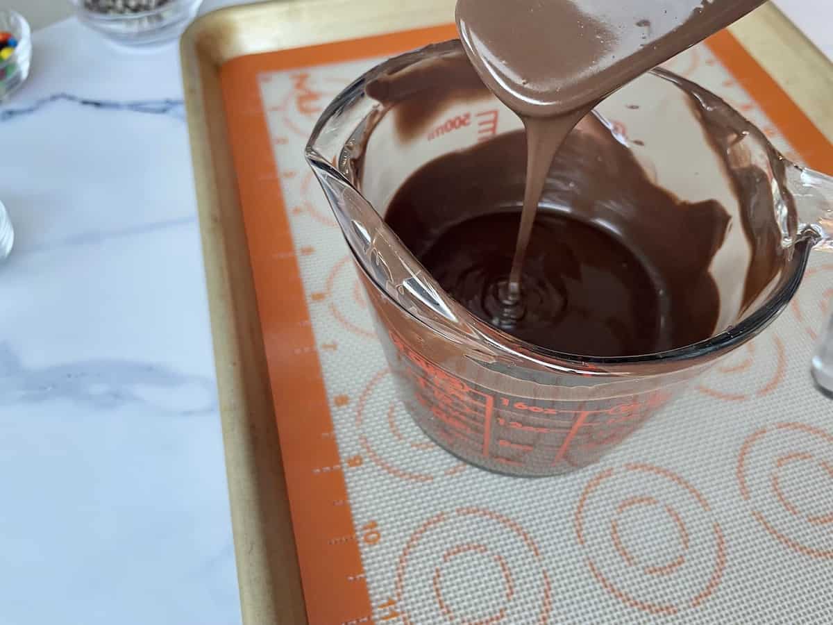 a 2 cup glass measuring cup with melted dark chocolate and a grey spatula with a cookie sheet and silpat in the background