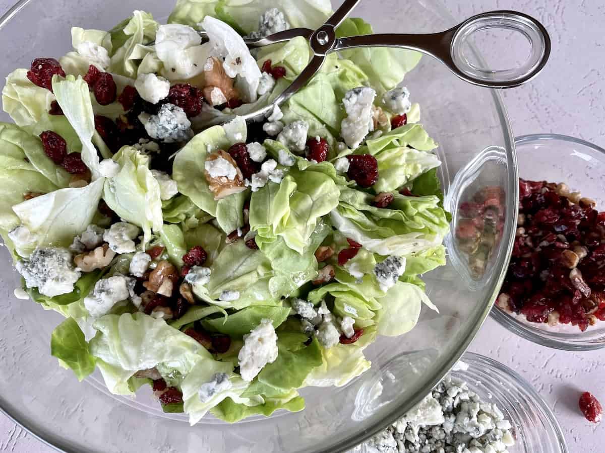 cranberry walnut and blue cheese salad in a glass bowl with silver serving tongs with additional toppings on the side