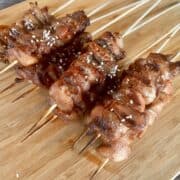 several skewers of cooked Chinese chicken on a stick resting on a wood cutting board garnished with sesame seeds