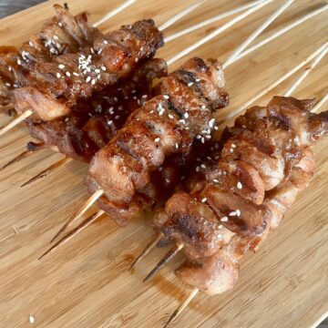 several skewers of cooked Chinese chicken on a stick resting on a wood cutting board garnished with sesame seeds
