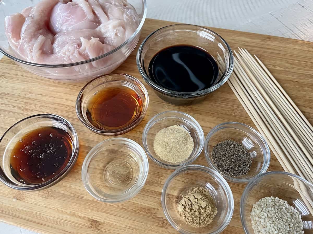 Chinese chicken ingredients layer out in small glass bowls with a pile of bamboo skewers on a wood cutting board