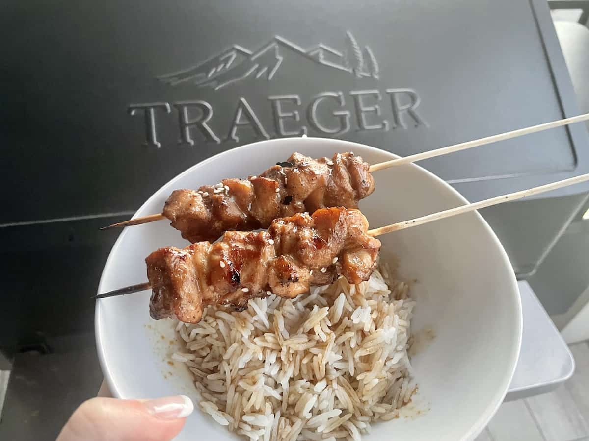 a bowl of white rice and two chicken skewers held up in front of a Traeger Grill 