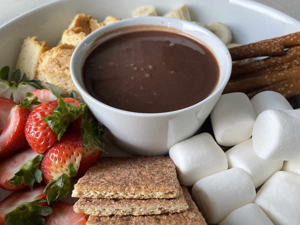 chocolate dipping sauce in a white porcelain bowl surrounded by cut bananas pound cake strawberries marshmallows and graham crackers