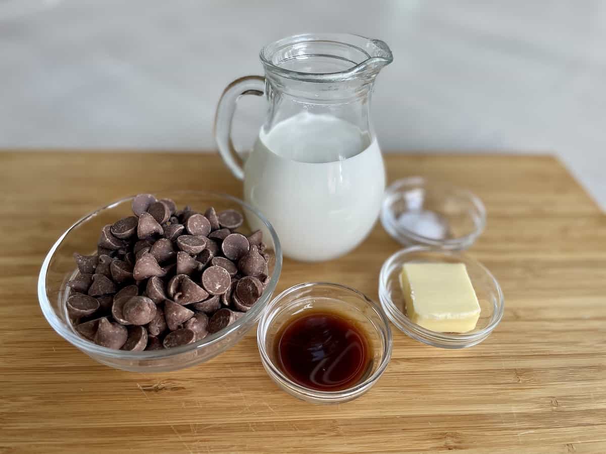 chocolate dipping sauce ingredients including a glass bowl of milk chocolate chips glass pitcher of heavy cream and small glass bowls of vanilla extract butter and salt