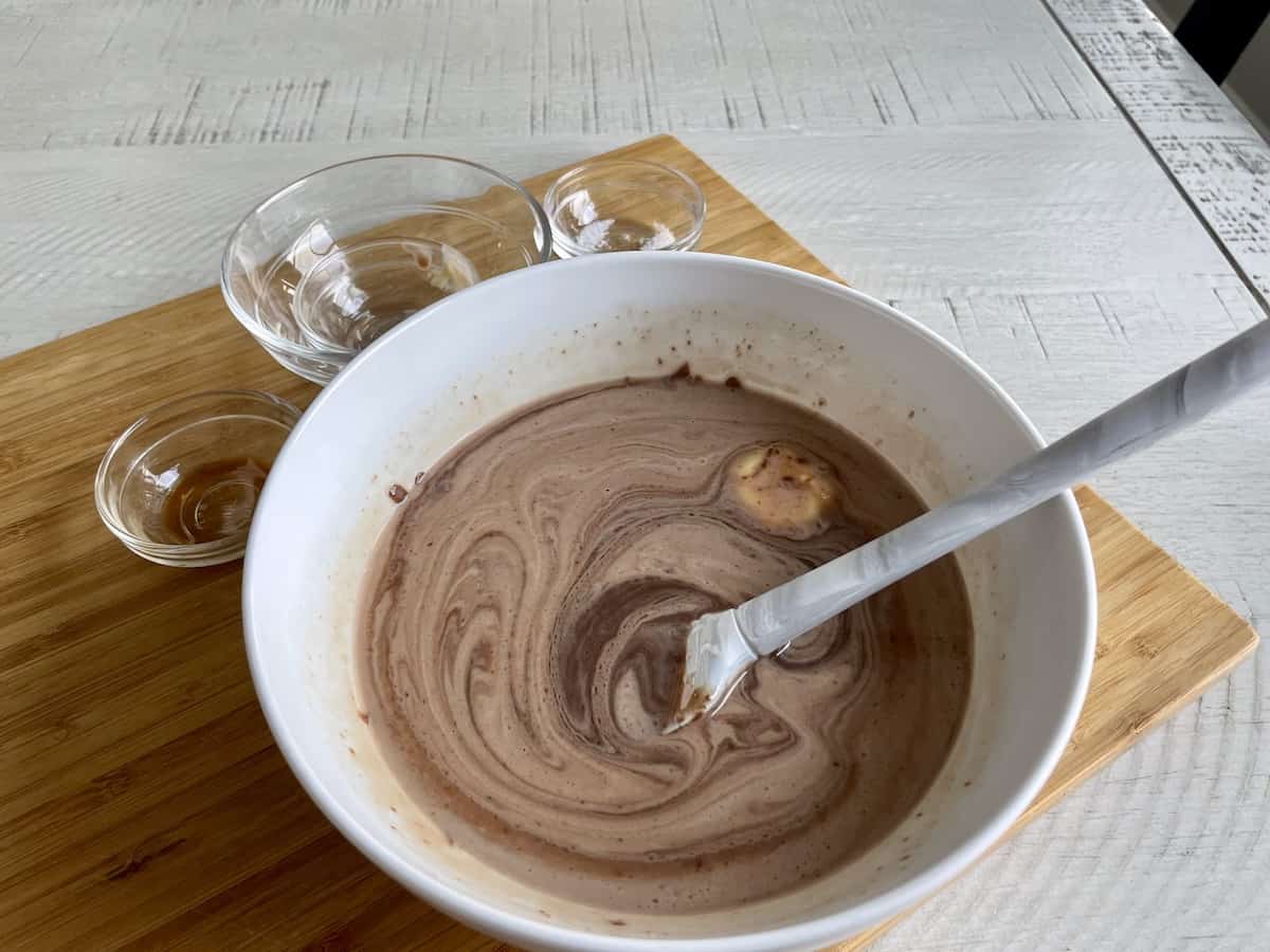 a white bowl of melted chocolate being stirred with a grey rubber spatula set on top of a wood cutting board