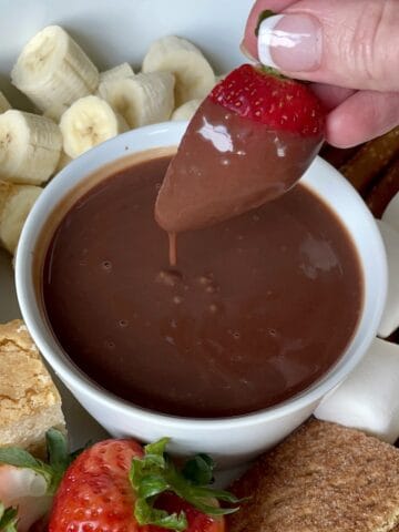 a strawberry being dipped into chocolate dipping sauce in a white porcelain bowl surrounded by cut bananas pound cake strawberries marshmallows and graham crackers