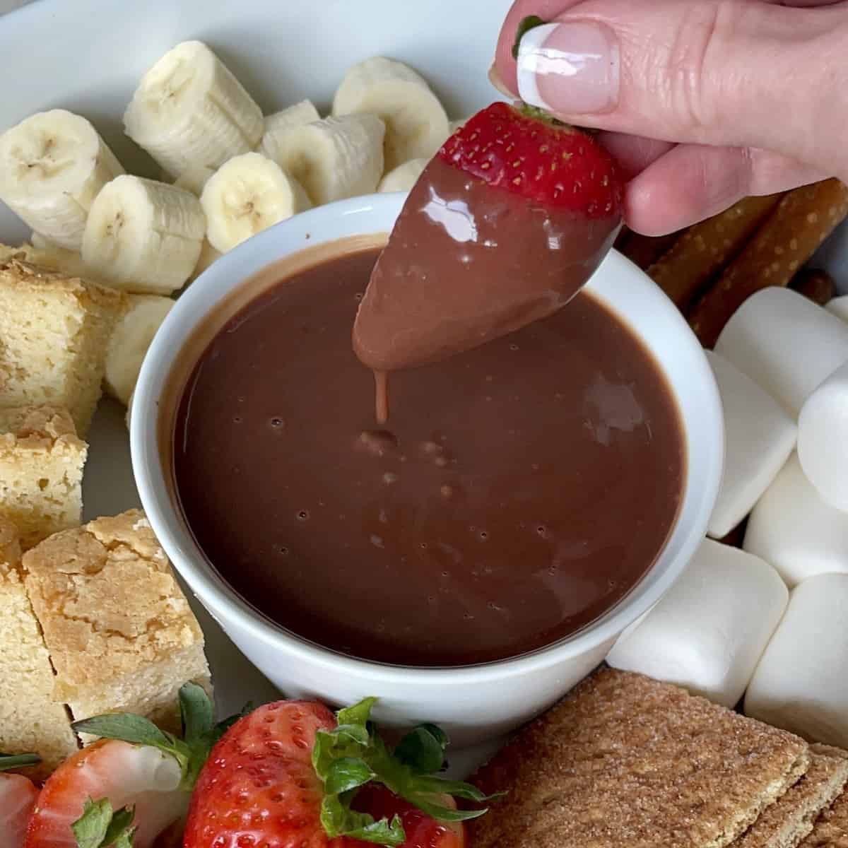 a strawberry being dipped into chocolate dipping sauce in a white porcelain bowl surrounded by cut bananas pound cake strawberries marshmallows and graham crackers