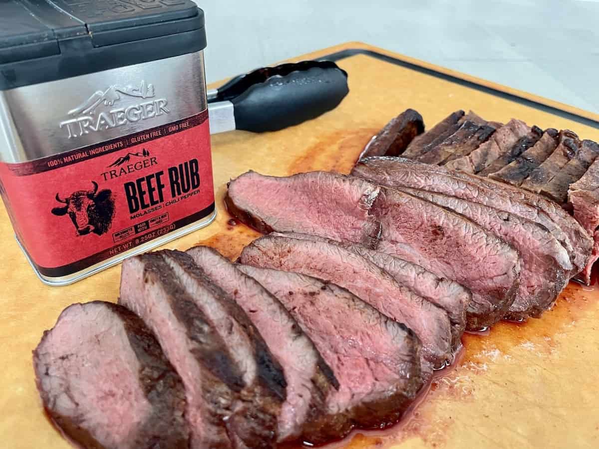 traeger tri tip sliced on a wood cutting board showing a dark brown outside and bright red inside with tongs and a container of traeger beef rub behind it

