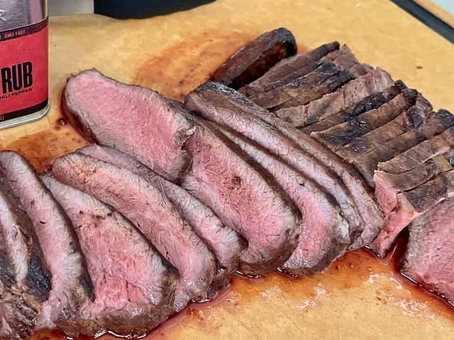 traeger tri tip sliced on a wood cutting board showing a dark brown outside and bright red inside with tongs and a container of traeger beef rub behind it