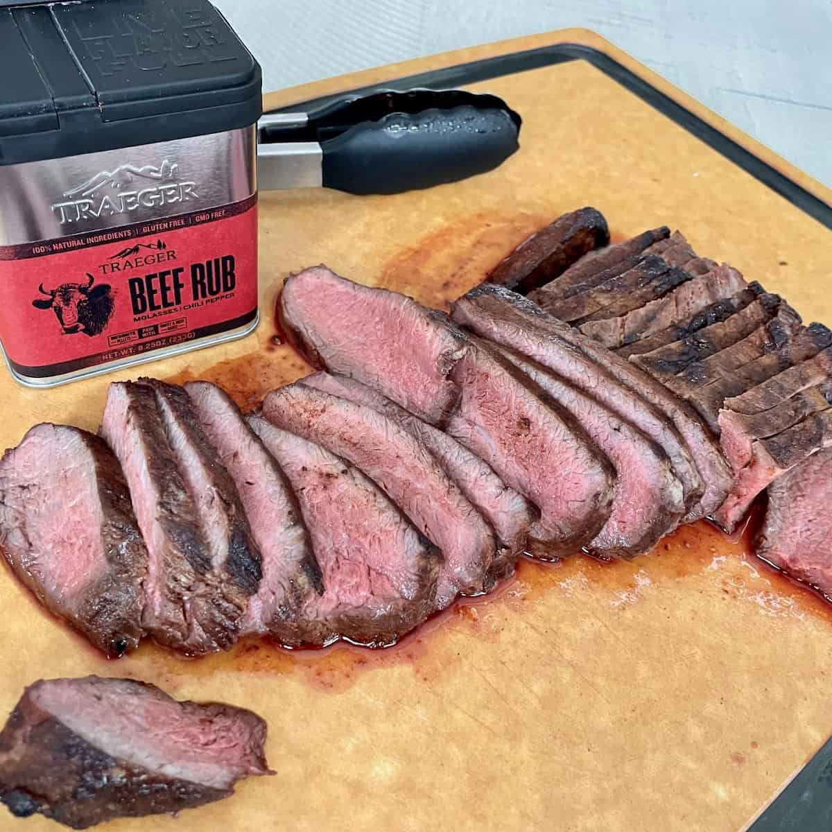 traeger tri tip sliced on a wood cutting board showing a dark brown outside and bright red inside with tongs and a container of traeger beef rub behind it