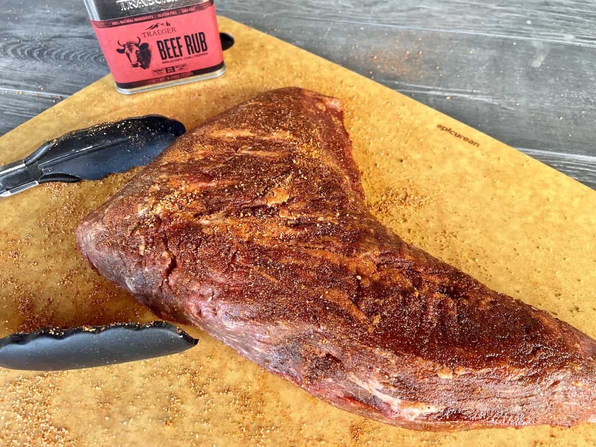 a raw tri tip covered in spices on a brown cutting board with black tongs next to ti and a container of traeger beef rub
