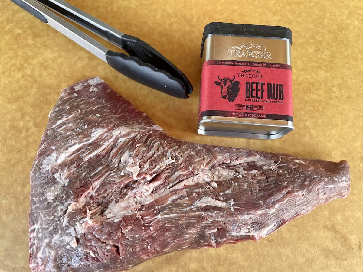 raw Tri tip on a brown cutting board with tongs and Traeger beef rub
