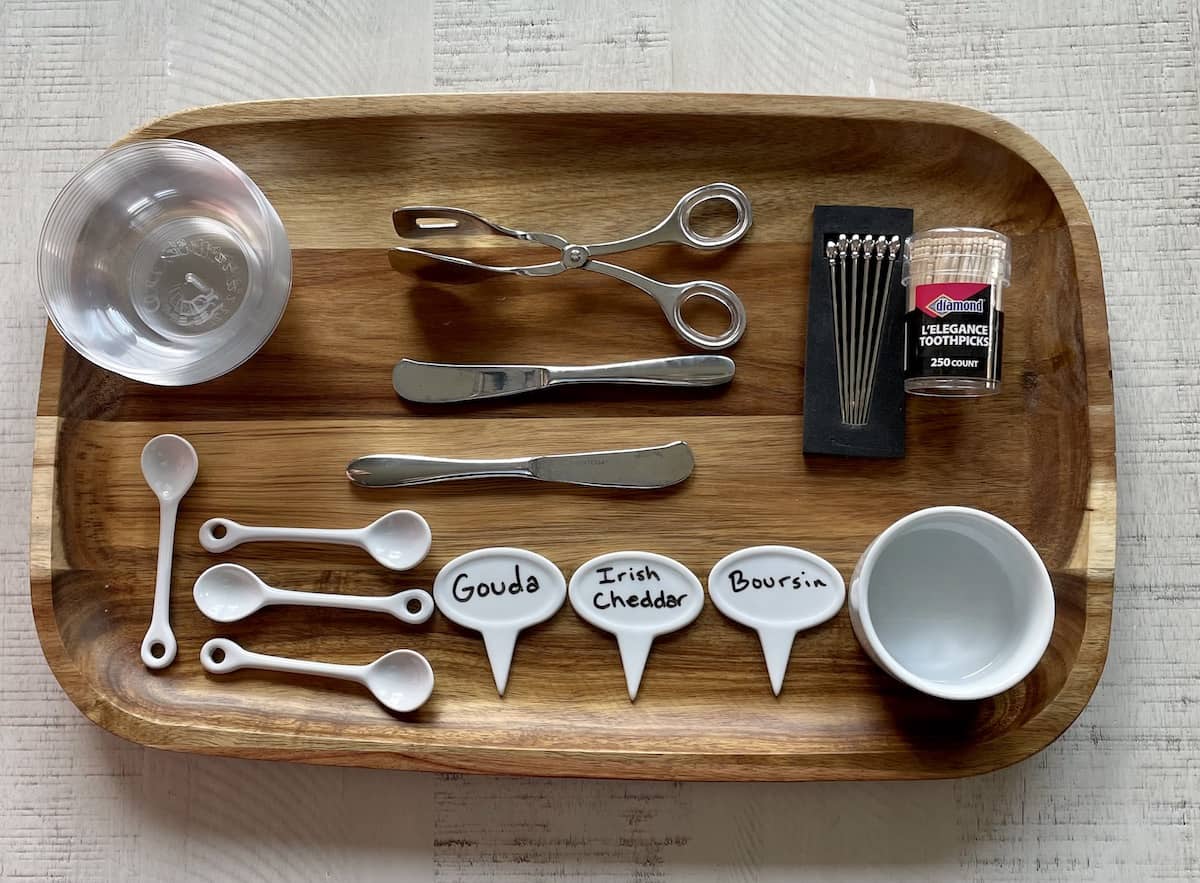 a wood board with small clear bowls small white serving spoons cheese markers tongs cheese knives toothpicks and a small white bowl on top