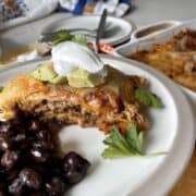 served piece of enchilada casserole on a white plate with black beans and garnished with cilantro avcado and sour cream