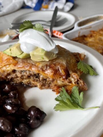 served piece of enchilada casserole on a white plate with black beans and garnished with cilantro avcado and sour cream