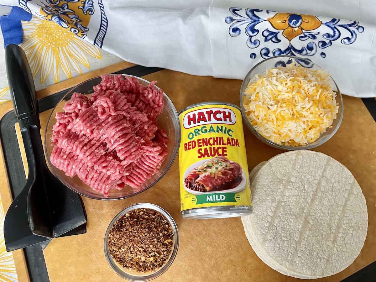 beef enchilada ingredients including raw ground beef in a glass bowl taco seasoning a can of hatch red enchilada sauce a pile of corn tortillas a bowl of cheese all laid out on a wood cutting board with an apron laying beside it
