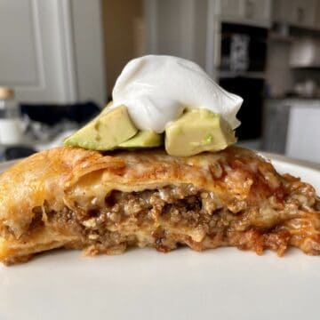 a piece of beef enchilada casserole on a white plate topped with diced avocado and a dollop of sour cream