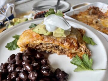 beef enchilada casserole served on a white plate with black beans diced avocado sour cream and cilantro with a casserole dish in the background