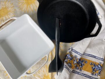 a white 9x9 baking dish meat masher and cast iron pan laid out on a table with a sunny tablecloth and yellow and blue apron