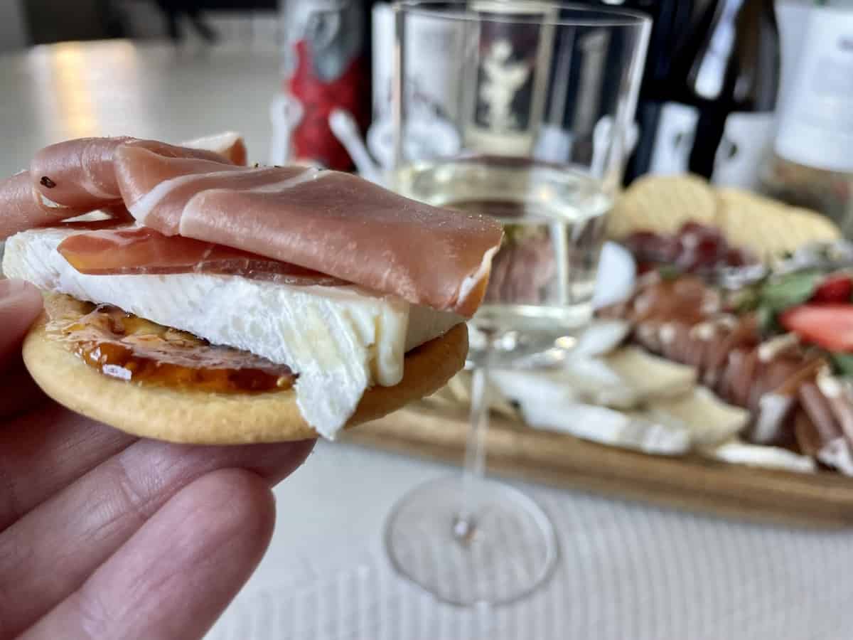 close up view of a cracker with fig spread brie cheese and prosciutto on top with a glass of pinot Grigio and more charcuterie and wine in the background