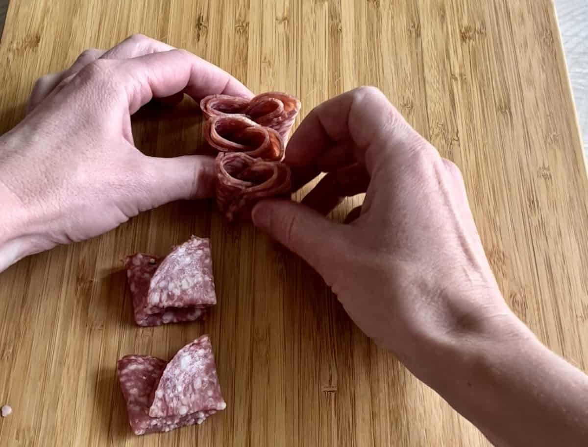 salami slices are being folded into quarters by two hands and placed together to create a salami train on a wood cutting board