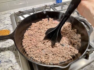 ground beef browning in a cast iron skillet with a meat masher breaking up the meat