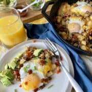 mexican breakfast skillet served on a white plate with avocado green onions and hot sauce served next to a cast iron skillet full of potatoes sausage cheese and eggs and garnishes in the background