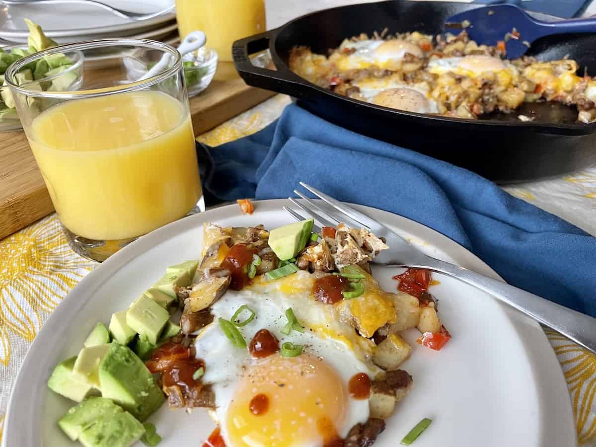 mexican breakfast skillet served on a white plate with avocado green onions and hot sauce served next to a cast iron skillet full of potatoes sausage cheese and eggs and garnishes in the background