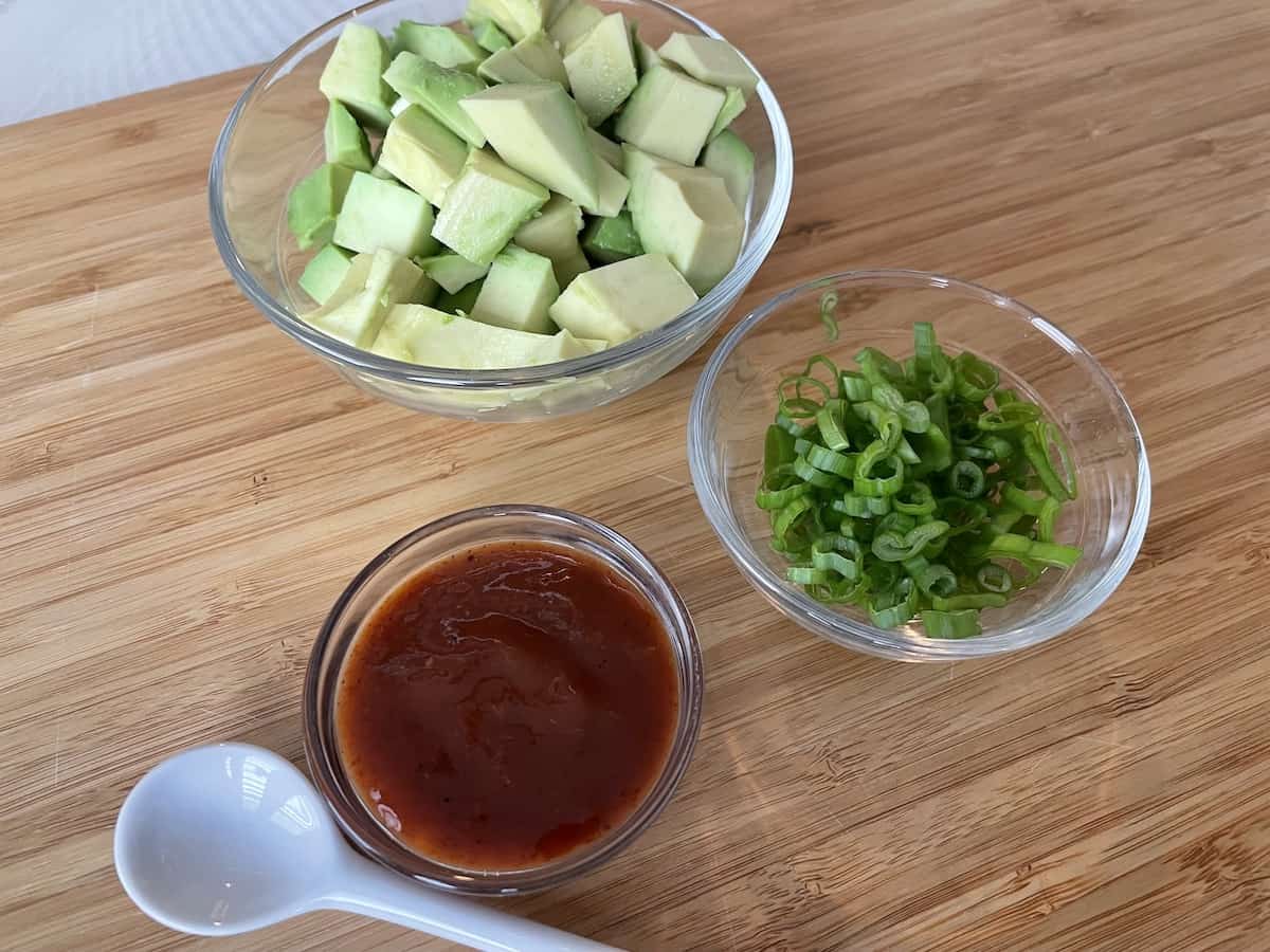 small glass bowls containing diced avocado diced green onions and hot sauce with a small white spoon