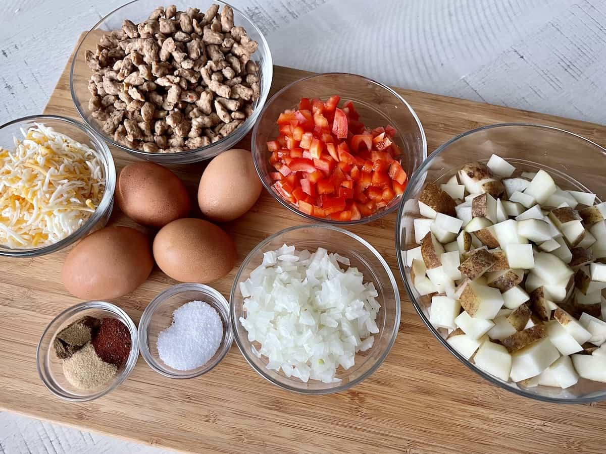 wood cutting board with ingredients including sausage eggs shredded cheese diced red bell pepper diced onion diced russet potatoes and spices