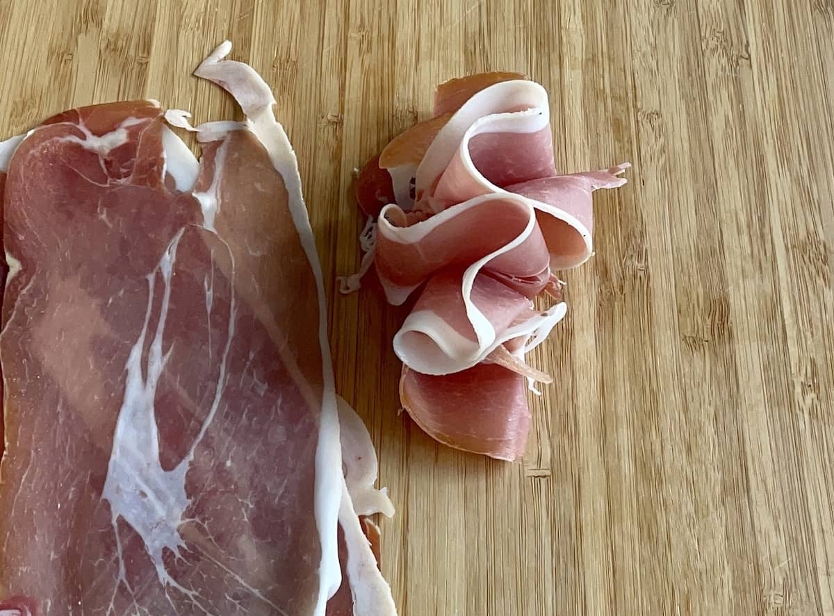 prosciutto made into ribbons by using an accordion fold and laid down on a wood cutting board with more prosciutto next to it