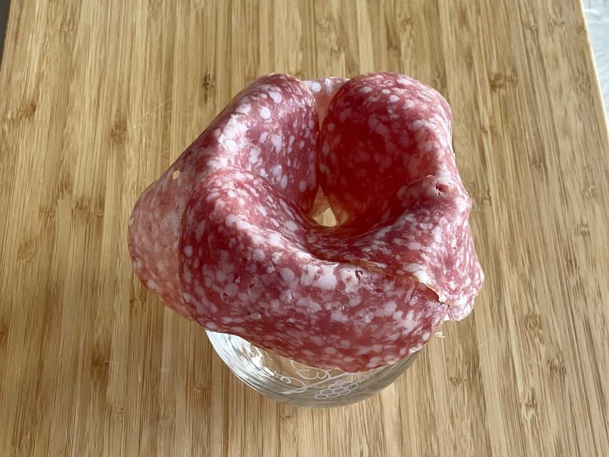 stemless wine glass on a wood cutting board with large salami folded over top of the glass opening