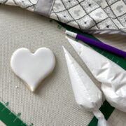 an iced heart shaped cookie on a white silpat with two bags of the best royal icing a cookie pick and a grey and white apron