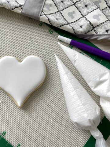 an iced heart shaped cookie on a white silpat with two bags of the best royal icing a cookie pick and a grey and white apron