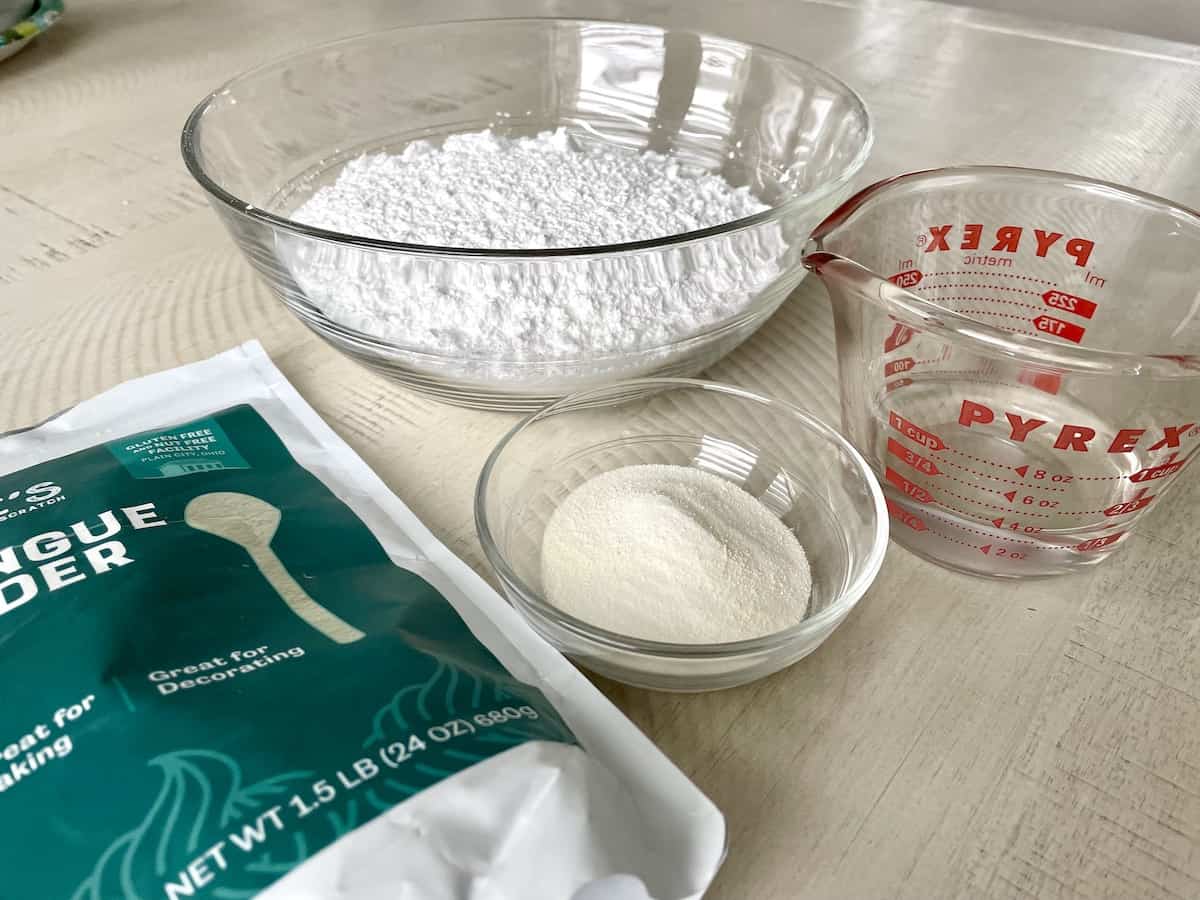 two glass bowls with powdered sugar meringue powder a glass measuring cup with water and a bag of judees meringue powder on a white kitchen table