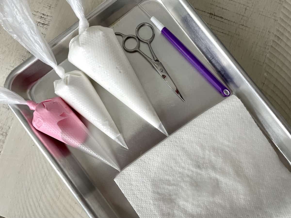 three piping bags of the best royal icing for sugar cookies on a metal tray with small scissors a cookie pick and a wet paper towel