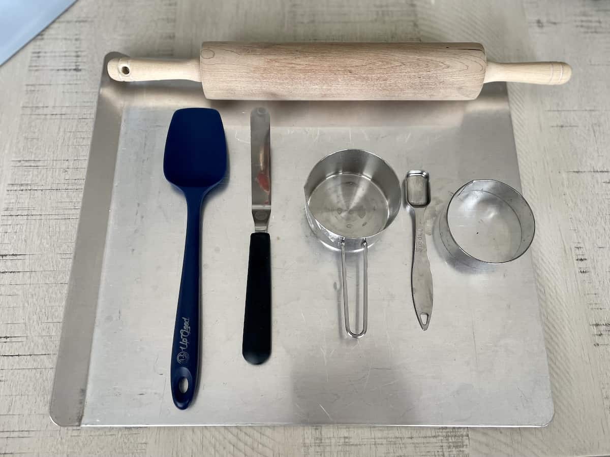 one baking sheet a blue rubber spatula a measuring cup rolling pin offset spatula a teaspoon and a cookie cutter