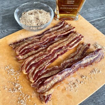 slices of bacon jerky on a wood cutting board with a bowl of brown sugar and bottle of angels envy bourbon