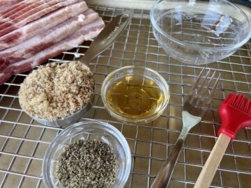 bacon jerky ingredients including thick cut bacon brown sugar bourbon in a glass bowl black pepper a fork pastry brush all set on a wire rack over a sheet pan
