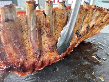 metal tongs holding up a rack of st Louis ribs on a black cutting board