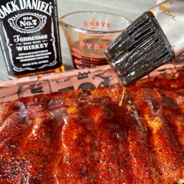 Jack Daniels sauce recipe in a measuring cup getting brushed onto cooked ribs with a bottle of Jack Daniels in the background