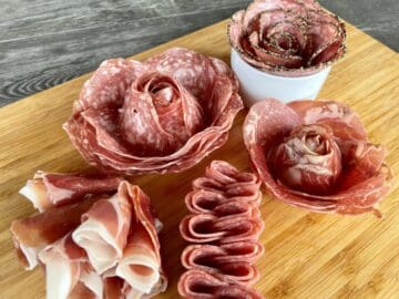 5 salami roses on a brown cutting board on a wood table