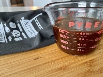 glass measuring cup with Jack Daniels sauce on a wood cutting board with a black grilling and bbq apron behind it