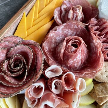 different sizes of meat roses on a charcuterie board with sliced apples crackers and cheddar cheese