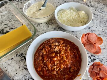 pizzagna recipe ingredients including cooked meat sauce no boil lasagna noodles ricotta cheese mixture shredded cheese pepperoni and parmesan next to a glass baking dish