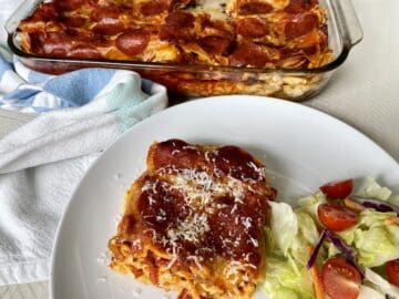 a slice of pizzagna on a white plate with a green salad and more pizzagna behind it