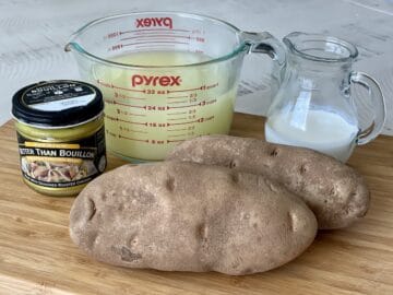 jar of better than bouillon with a glass measuring cup of stock next to two russet potatoes and a glass pitcher of heavy cream