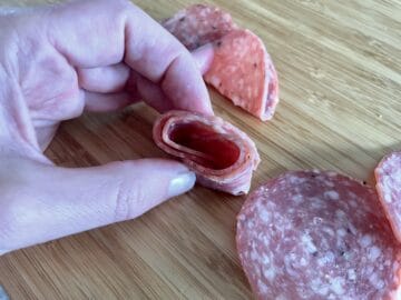a hand holding two salami slices folded in half and then folded around each other on a wood cutting board